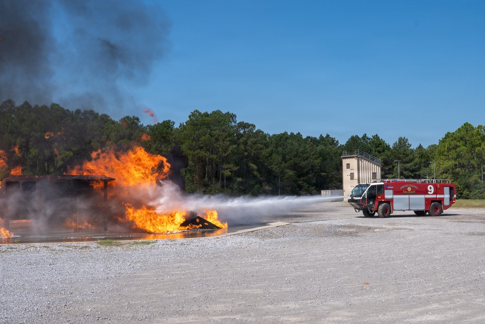 A United States Air Force fire truck (numbered 9) is actively discharging a water/foam stream at a large controlled fire during what appears to be a firefighting training exercise. The fire is burning intensely on a flat surface, producing large orange flames and thick black smoke. The setting is an open gravel training area with a concrete observation or control tower visible in the background.