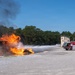 A United States Air Force fire truck (numbered 9) is actively discharging a water/foam stream at a large controlled fire during what appears to be a firefighting training exercise. The fire is burning intensely on a flat surface, producing large orange flames and thick black smoke. The setting is an open gravel training area with a concrete observation or control tower visible in the background.