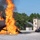 Gulfport CRTC Live Fire Training Exercise A fire training exercise is underway at what appears to be a military or emergency services training facility. A large, intense orange flame erupts from a metal aircraft fuselage mockup on a gravel pad, while a red fire truck directs a water stream toward the blaze. The controlled environment, purpose-built structures, and training props indicate this is a live-fire firefighting drill.