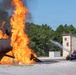 A fire training exercise is underway at what appears to be a military or emergency services training facility. A large, intense orange flame erupts from a metal aircraft fuselage mockup on a gravel pad, while a red fire truck directs a water stream toward the blaze. The controlled environment, purpose-built structures, and training props indicate this is a live-fire firefighting drill.