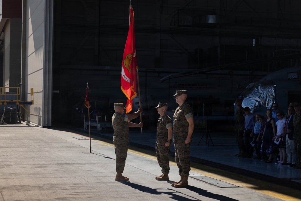 A U.S. Marine Corps change of command ceremony is taking place on an aircraft hangar tarmac. Three Marines in woodland MARPAT camouflage uniforms are participating in the formal guidon transfer, with one Marine holding a red unit colors flag bearing the Marine Corps emblem. Civilian spectators and additional military personnel are visible in the background inside the hangar.
