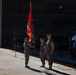 A U.S. Marine Corps change of command ceremony is taking place on an aircraft hangar tarmac. Three Marines in woodland MARPAT camouflage uniforms are participating in the formal guidon transfer, with one Marine holding a red unit colors flag bearing the Marine Corps emblem. Civilian spectators and additional military personnel are visible in the background inside the hangar.