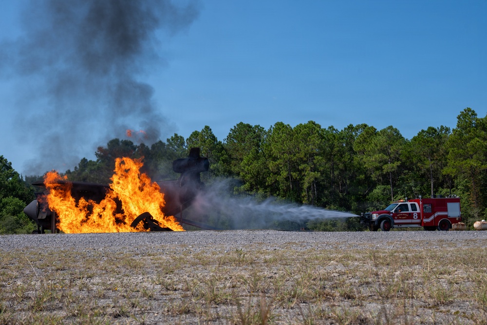 A red fire truck (Unit 8) is actively spraying water or foam onto a large, intensely burning fire on a gravel surface. The fire appears to involve a metal structure or aircraft mockup, producing tall orange flames and thick black smoke. This appears to be a controlled firefighting training exercise conducted in an open area bordered by pine trees.