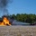 Gulfport CRTC Live Fire Training Exercise A red fire truck (Unit 8) is actively spraying water or foam onto a large, intensely burning fire on a gravel surface. The fire appears to involve a metal structure or aircraft mockup, producing tall orange flames and thick black smoke. This appears to be a controlled firefighting training exercise conducted in an open area bordered by pine trees.