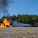 A red fire truck (Unit 8) is actively spraying water or foam onto a large, intensely burning fire on a gravel surface. The fire appears to involve a metal structure or aircraft mockup, producing tall orange flames and thick black smoke. This appears to be a controlled firefighting training exercise conducted in an open area bordered by pine trees.