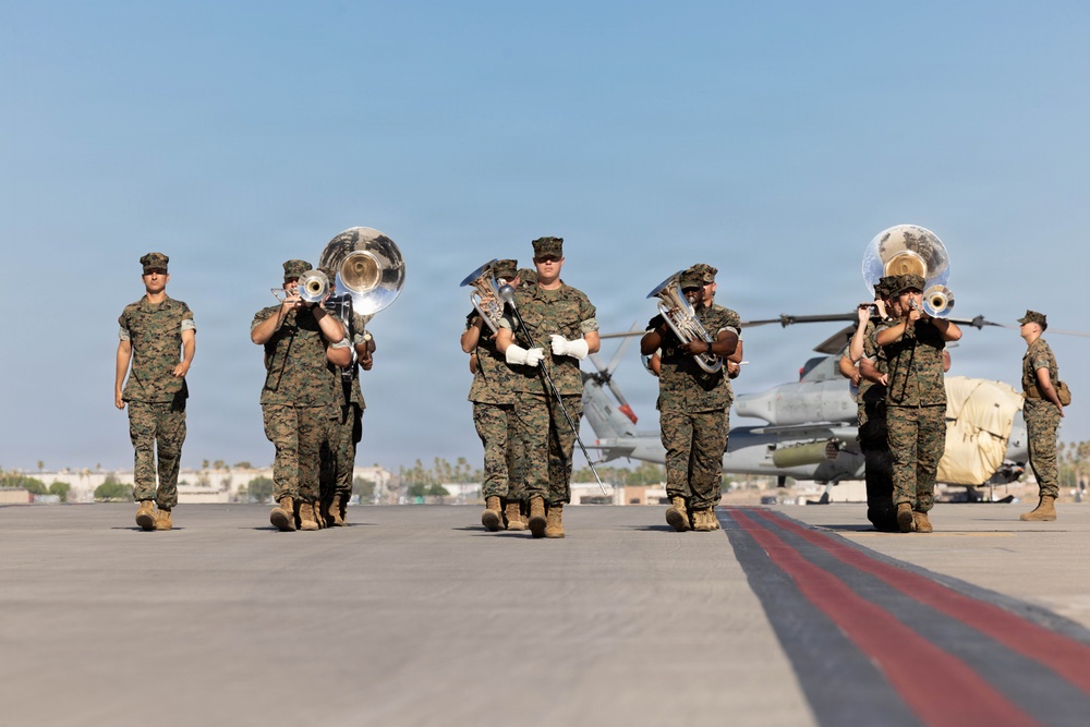A U.S. Marine Corps marching band performs on a military airfield tarmac, with musicians in woodland MARPAT camouflage uniforms playing brass instruments. The band is marching in formation across the flight line, with military aircraft visible in the background. The setting appears to be a formal ceremony or parade at a Marine Corps air station.