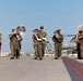 A U.S. Marine Corps marching band performs on a military airfield tarmac, with musicians in woodland MARPAT camouflage uniforms playing brass instruments. The band is marching in formation across the flight line, with military aircraft visible in the background. The setting appears to be a formal ceremony or parade at a Marine Corps air station.