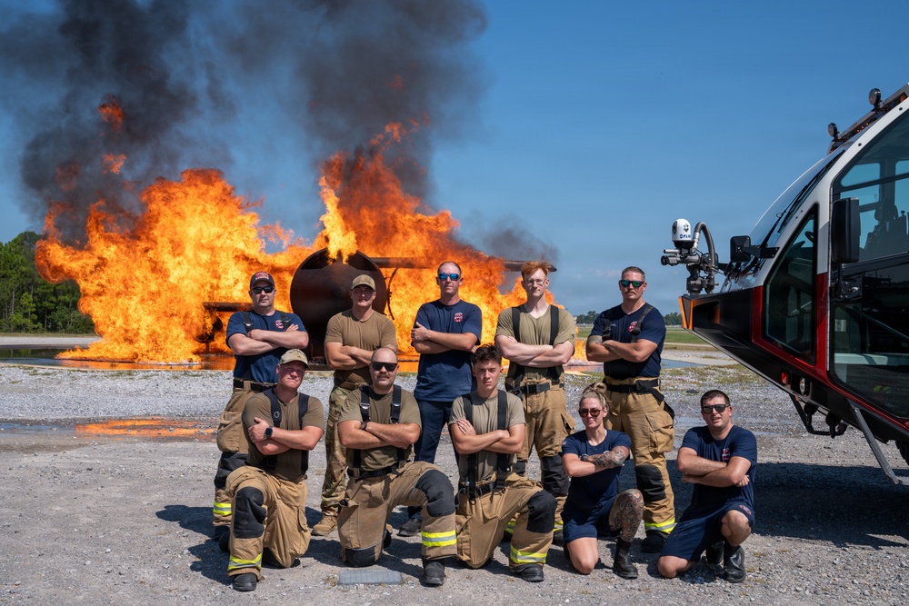 A group of approximately ten firefighters pose confidently in front of a large controlled burn, likely during a training exercise. The team is arranged in two rows — standing and kneeling — with arms crossed in a formal group photo stance. A specialized fire/rescue vehicle is visible on the right side of the frame.