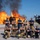 Gulfport CRTC Live Fire Training Exercise A group of approximately ten firefighters pose confidently in front of a large controlled burn, likely during a training exercise. The team is arranged in two rows — standing and kneeling — with arms crossed in a formal group photo stance. A specialized fire/rescue vehicle is visible on the right side of the frame.