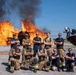 A group of approximately ten firefighters pose confidently in front of a large controlled burn, likely during a training exercise. The team is arranged in two rows — standing and kneeling — with arms crossed in a formal group photo stance. A specialized fire/rescue vehicle is visible on the right side of the frame.