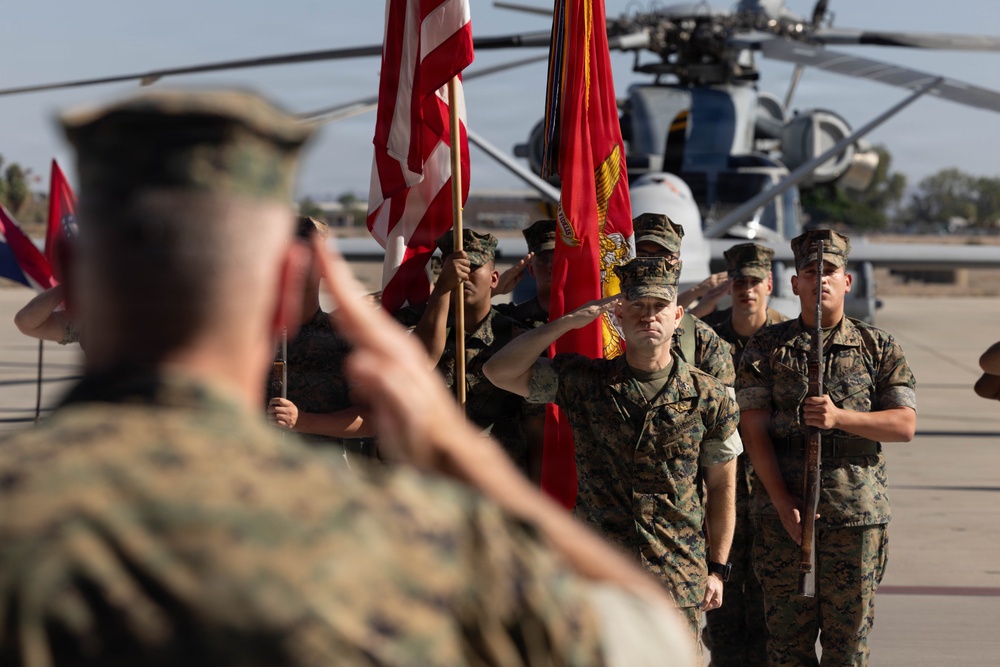 A formal U.S. Marine Corps military ceremony is taking place on an airfield tarmac. Personnel in MARPAT camouflage uniforms are rendering salutes while a color guard holds the U.S. flag and Marine Corps colors. A military helicopter is visible in the background, suggesting the ceremony is at a Marine Corps air station.