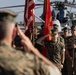 A formal U.S. Marine Corps military ceremony is taking place on an airfield tarmac. Personnel in MARPAT camouflage uniforms are rendering salutes while a color guard holds the U.S. flag and Marine Corps colors. A military helicopter is visible in the background, suggesting the ceremony is at a Marine Corps air station.