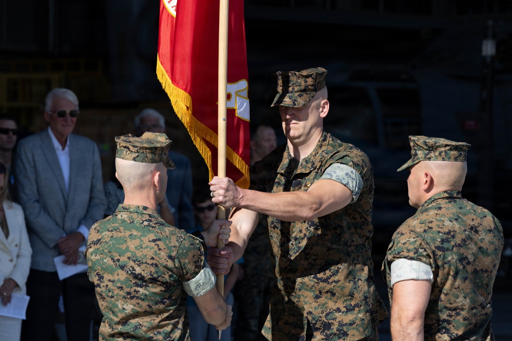 A U.S. Marine Corps change of command ceremony is taking place, with two Marines transferring a red unit guidon flag between them. Three Marines in MARPAT woodland camouflage uniforms are the central figures, while civilian observers watch in the background. The passing of the colors is a formal military tradition symbolizing the transfer of command authority.