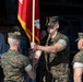 A U.S. Marine Corps change of command ceremony is taking place, with two Marines transferring a red unit guidon flag between them. Three Marines in MARPAT woodland camouflage uniforms are the central figures, while civilian observers watch in the background. The passing of the colors is a formal military tradition symbolizing the transfer of command authority.