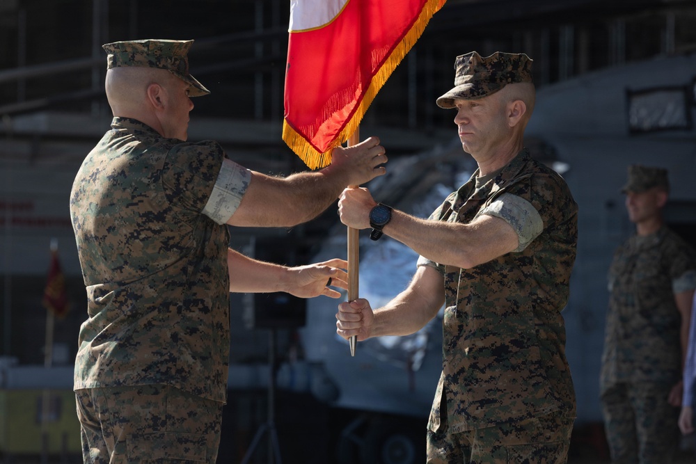 Two U.S. Marine Corps officers are participating in a formal change of command ceremony, passing a guidon (unit flag) between them. The red and gold flag with gold fringe is characteristic of Marine Corps unit colors. Additional Marines in uniform are visible in the background, consistent with a formal military ceremony setting.