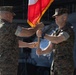 Two U.S. Marine Corps officers are participating in a formal change of command ceremony, passing a guidon (unit flag) between them. The red and gold flag with gold fringe is characteristic of Marine Corps unit colors. Additional Marines in uniform are visible in the background, consistent with a formal military ceremony setting.