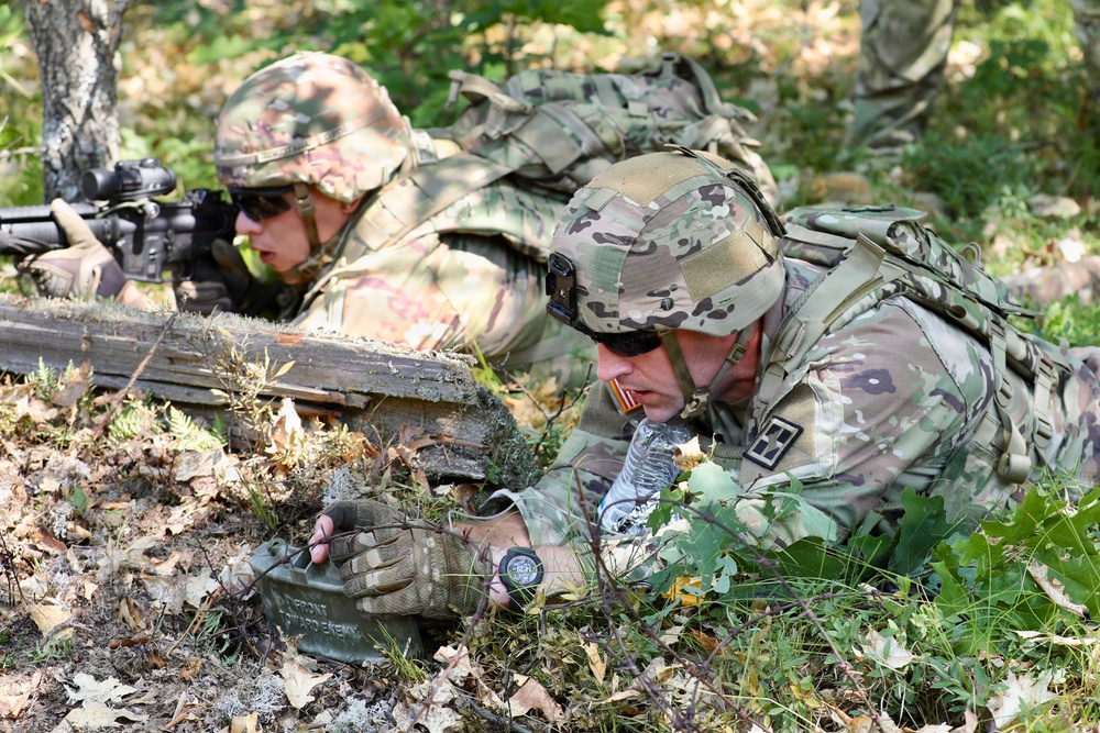 Two soldiers in full camouflage gear are in a prone position in a wooded area, appearing to be conducting a tactical field exercise. The foreground soldier is examining or handling a Claymore mine, while the background soldier is aiming a rifle. Both wear MultiCam pattern uniforms with tactical helmets, body armor, and rucksacks.
