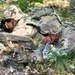 Two soldiers in full camouflage gear are in a prone position in a wooded area, appearing to be conducting a tactical field exercise. The foreground soldier is examining or handling a Claymore mine, while the background soldier is aiming a rifle. Both wear MultiCam pattern uniforms with tactical helmets, body armor, and rucksacks.