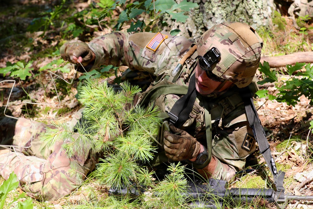 A U.S. Army soldier in full combat gear is low-crawling through a wooded forest environment. The soldier is wearing Operational Camouflage Pattern (OCP) uniform with a U.S. flag patch visible on the left shoulder. The soldier appears to be conducting a field training exercise, moving tactically through dense undergrowth.