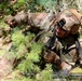 A U.S. Army soldier in full combat gear is low-crawling through a wooded forest environment. The soldier is wearing Operational Camouflage Pattern (OCP) uniform with a U.S. flag patch visible on the left shoulder. The soldier appears to be conducting a field training exercise, moving tactically through dense undergrowth.