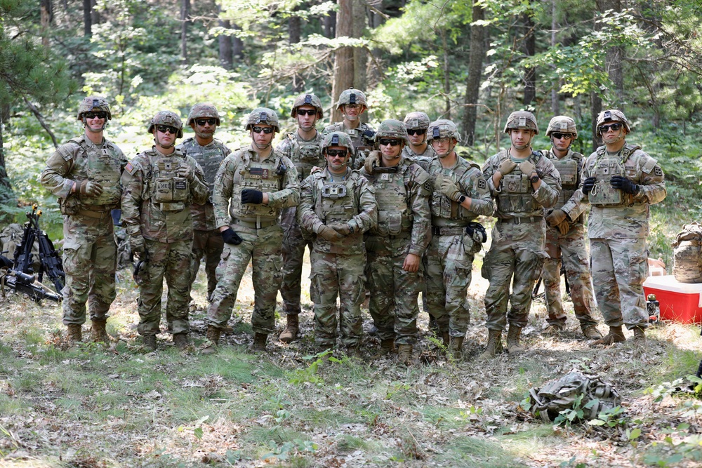A group of approximately 13 military personnel in U.S. Army MultiCam (OCP) camouflage uniforms pose together in a wooded outdoor setting. They are wearing combat helmets, tactical vests, and gloves, suggesting a field training exercise or operational environment. The group appears relaxed and is posing for a group photograph.