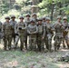A group of approximately 13 military personnel in U.S. Army MultiCam (OCP) camouflage uniforms pose together in a wooded outdoor setting. They are wearing combat helmets, tactical vests, and gloves, suggesting a field training exercise or operational environment. The group appears relaxed and is posing for a group photograph.
