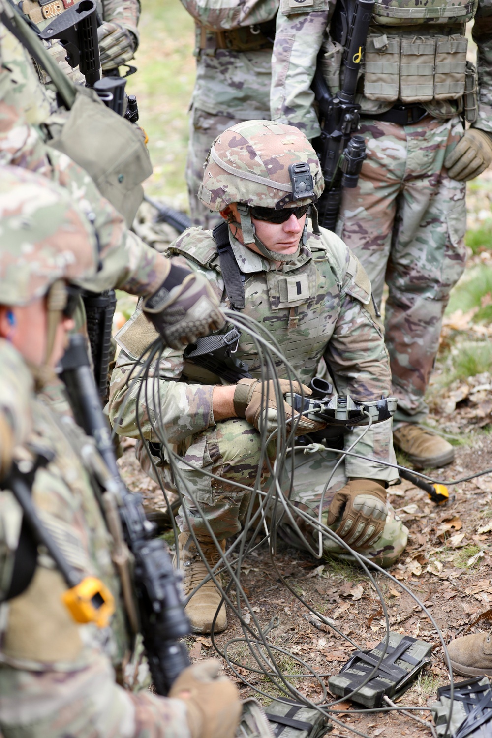 A group of military personnel in Operational Camouflage Pattern (OCP) uniforms are gathered in a wooded outdoor setting. The central figure is kneeling on the ground, handling what appears to be a coil of wire or cable connected to a device, possibly conducting demolitions, communications, or IED-related training. Surrounding soldiers are equipped with rifles, tactical vests, and helmets.