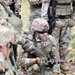 A group of military personnel in Operational Camouflage Pattern (OCP) uniforms are gathered in a wooded outdoor setting. The central figure is kneeling on the ground, handling what appears to be a coil of wire or cable connected to a device, possibly conducting demolitions, communications, or IED-related training. Surrounding soldiers are equipped with rifles, tactical vests, and helmets.