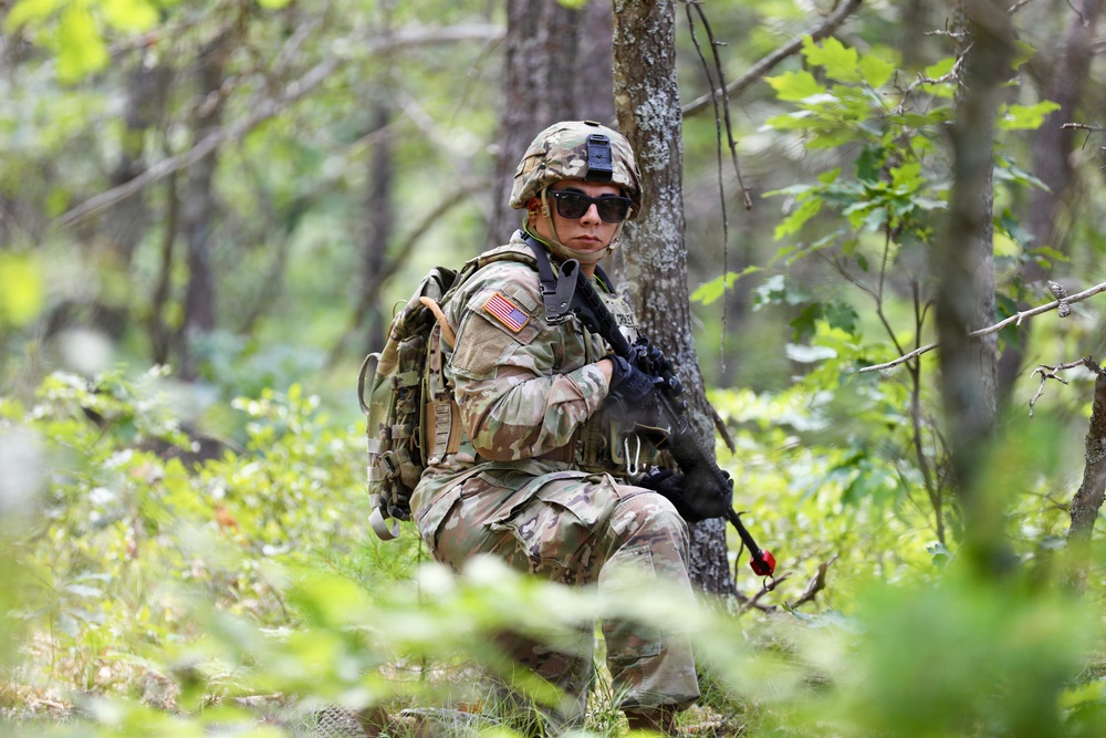 A U.S. Army soldier in full combat gear is positioned in a wooded area, appearing to be on patrol or participating in a field training exercise. The soldier is wearing Operational Camouflage Pattern (OCP) uniform with a visible American flag patch on the right shoulder, tactical vest, helmet, and sunglasses. The soldier is crouched near a tree while holding a rifle, surrounded by dense green forest vegetation.