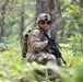 A U.S. Army soldier in full combat gear is positioned in a wooded area, appearing to be on patrol or participating in a field training exercise. The soldier is wearing Operational Camouflage Pattern (OCP) uniform with a visible American flag patch on the right shoulder, tactical vest, helmet, and sunglasses. The soldier is crouched near a tree while holding a rifle, surrounded by dense green forest vegetation.