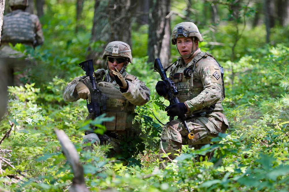 Two soldiers in U.S. Army MultiCam (OCP) uniforms are positioned in a wooded area, appearing to communicate and coordinate during what looks like a field training exercise. Both are equipped with tactical gear, helmets, plate carriers, and rifles. A third soldier is partially visible in the background.