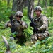 Two soldiers in U.S. Army MultiCam (OCP) uniforms are positioned in a wooded area, appearing to communicate and coordinate during what looks like a field training exercise. Both are equipped with tactical gear, helmets, plate carriers, and rifles. A third soldier is partially visible in the background.