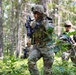 The image shows multiple soldiers in full combat gear moving through a forested area during what appears to be a military training exercise. They are wearing OCP (Operational Camouflage Pattern) uniforms and carrying assault rifles while navigating through dense woodland undergrowth. The scene suggests a patrol or tactical movement drill in a temperate forest environment.