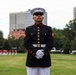 A United States Marine in full dress blues uniform stands at parade rest on a grass field, likely during a formal ceremony or parade. The Marine wears the distinctive black and red dress uniform with gold buttons, white gloves, white belt, and white trousers. In the background, additional Marines in red uniforms are visible, along with urban buildings and trees.