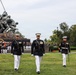 Four U.S. Marine Corps officers in dress blue uniforms march in formation at the Marine Corps War Memorial (Iwo Jima Memorial) in Arlington, Virginia. Behind them, a large formation of Marines in dress whites and blues stands at attention near the iconic bronze statue depicting the flag-raising at Iwo Jima. This appears to be a formal ceremonial parade or sunset parade event.