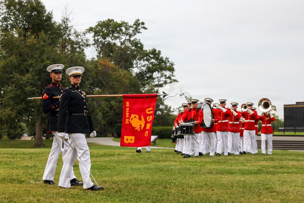 A U.S. Marine Corps Band (USMC B) from Washington, DC is performing a ceremonial parade or drill on a grass field. Two Marines in dress blues with white trousers lead the formation, one carrying a red guidon flag bearing 'USMC B' and 'HQ WASH DC' with the Marine Corps eagle, globe, and anchor emblem. Behind them, band members in red tunics and white trousers play brass and percussion instruments.