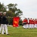 A U.S. Marine Corps Band (USMC B) from Washington, DC is performing a ceremonial parade or drill on a grass field. Two Marines in dress blues with white trousers lead the formation, one carrying a red guidon flag bearing 'USMC B' and 'HQ WASH DC' with the Marine Corps eagle, globe, and anchor emblem. Behind them, band members in red tunics and white trousers play brass and percussion instruments.