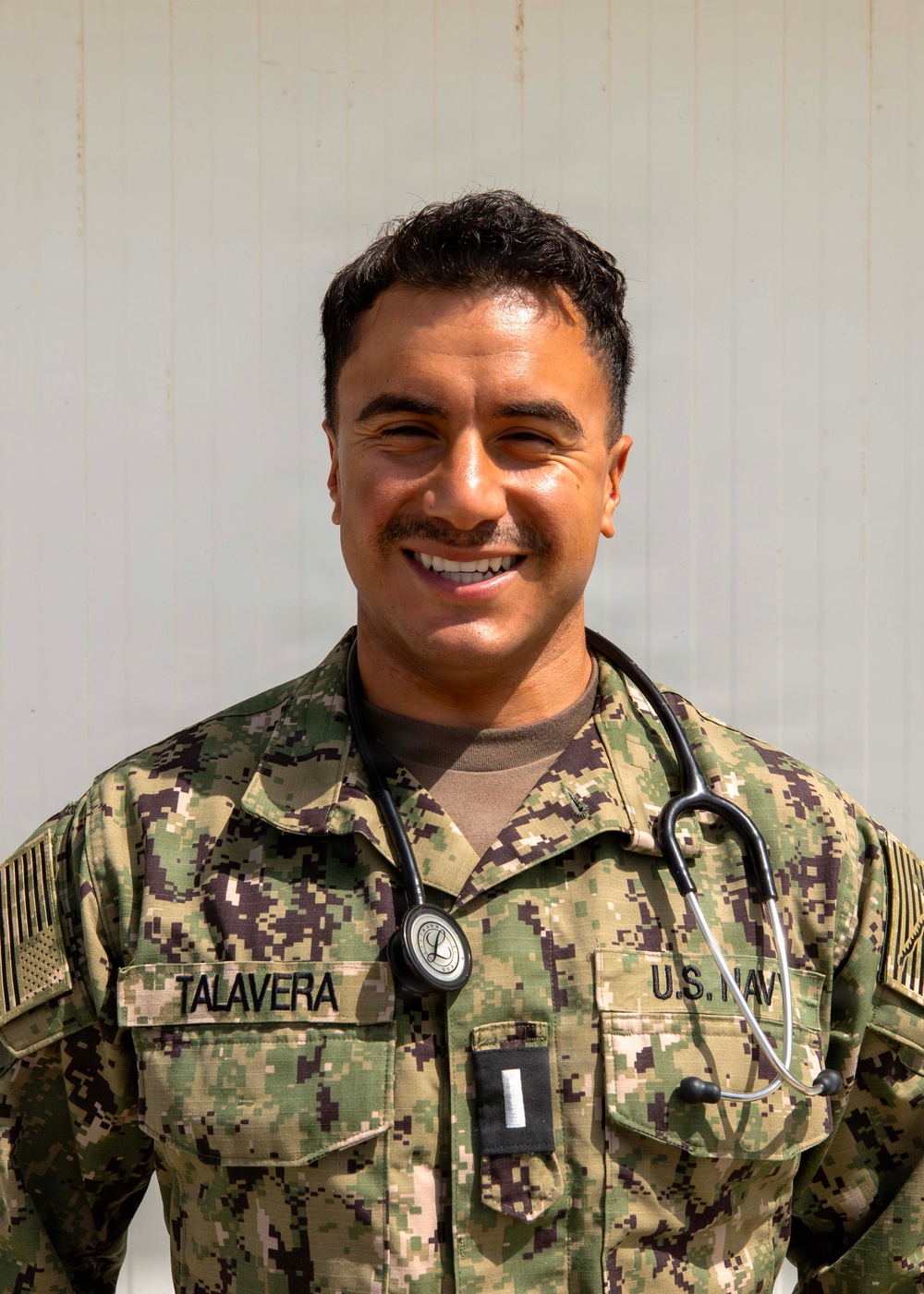 A U.S. Navy service member named Talavera is photographed in a formal portrait-style image. The individual is wearing Navy Working Uniform (NWU) Type III camouflage and has a stethoscope around their neck, indicating a medical role. A rank insignia visible on the chest pocket suggests the rank of Ensign (O-1).
