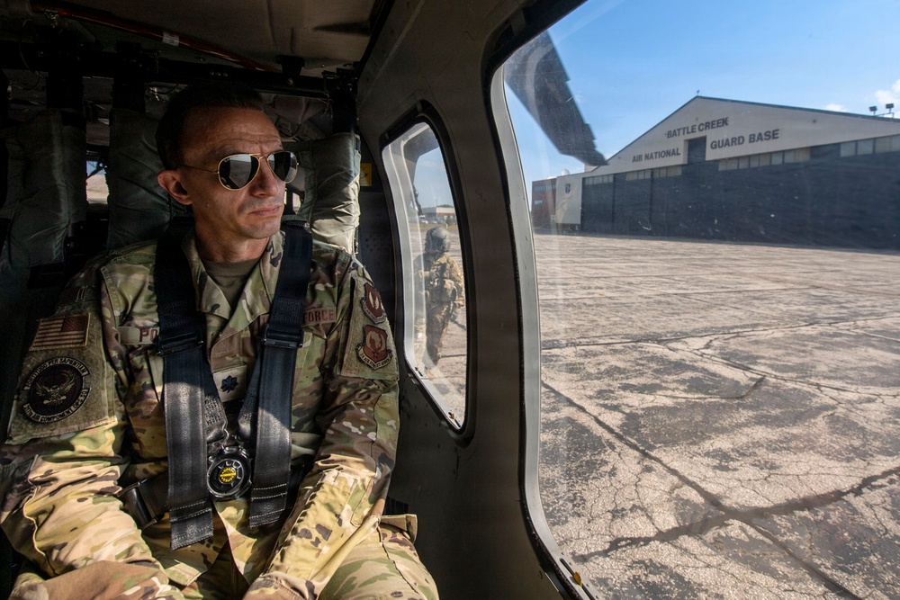 A military personnel member in US Air Force camouflage uniform is seated inside a helicopter, secured with a harness/seatbelt system and wearing aviator sunglasses. Through the helicopter window, the Battle Creek Air National Guard Base facility and tarmac are clearly visible. The image appears to be taken during a flight operation or ground taxi at the base.