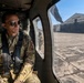 A military personnel member in US Air Force camouflage uniform is seated inside a helicopter, secured with a harness/seatbelt system and wearing aviator sunglasses. Through the helicopter window, the Battle Creek Air National Guard Base facility and tarmac are clearly visible. The image appears to be taken during a flight operation or ground taxi at the base.