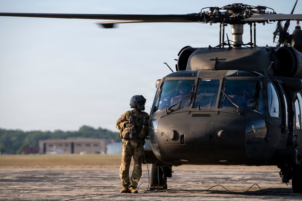 A military personnel in full combat gear stands on a tarmac facing a large dark-colored military helicopter, appearing to communicate with the cockpit crew. The helicopter, identifiable as a UH-60 Black Hawk, is positioned on what appears to be a military airfield with trees and buildings visible in the background. The image is taken during daylight hours with warm, low-angle lighting suggesting early morning or late afternoon conditions.