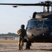 A military personnel in full combat gear stands on a tarmac facing a large dark-colored military helicopter, appearing to communicate with the cockpit crew. The helicopter, identifiable as a UH-60 Black Hawk, is positioned on what appears to be a military airfield with trees and buildings visible in the background. The image is taken during daylight hours with warm, low-angle lighting suggesting early morning or late afternoon conditions.
