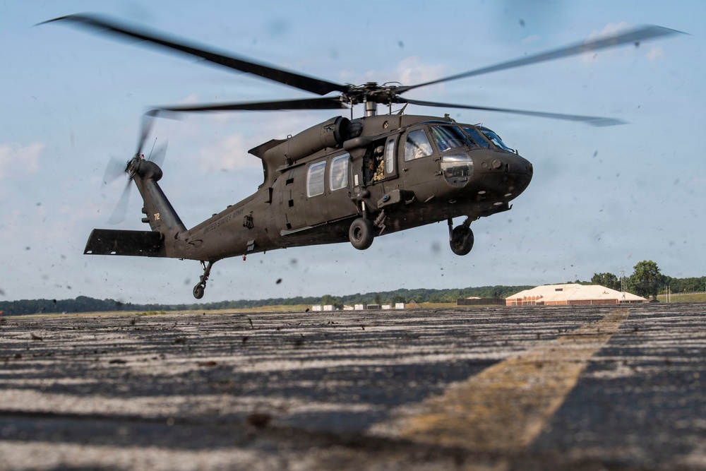 A U.S. Army UH-60 Black Hawk helicopter is captured in a low hover or takeoff phase above a tarmac or airfield surface. The rotor wash is visibly kicking up debris from the ground surface. A hangar building and tree line are visible in the background.