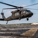 A U.S. Army UH-60 Black Hawk helicopter is captured in a low hover or takeoff phase above a tarmac or airfield surface. The rotor wash is visibly kicking up debris from the ground surface. A hangar building and tree line are visible in the background.