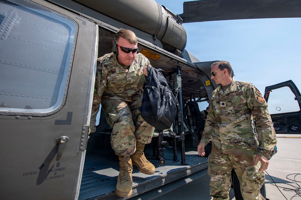 Two military personnel in OCP (Operational Camouflage Pattern) uniforms are photographed near a military helicopter, likely a UH-60 Black Hawk. One soldier is exiting or stepping out of the aircraft's side door while carrying a black backpack, while the second stands nearby on the tarmac. The setting appears to be a military airfield or flight line under clear skies.