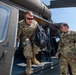 Two military personnel in OCP (Operational Camouflage Pattern) uniforms are photographed near a military helicopter, likely a UH-60 Black Hawk. One soldier is exiting or stepping out of the aircraft's side door while carrying a black backpack, while the second stands nearby on the tarmac. The setting appears to be a military airfield or flight line under clear skies.