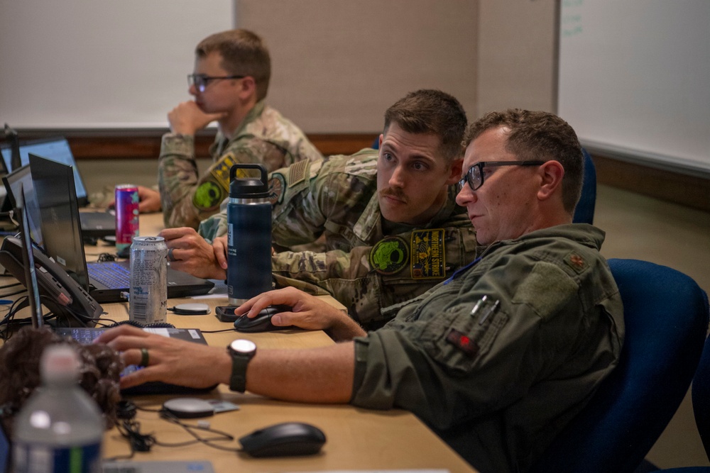 Three military personnel in camouflage and flight suits are working at a desk with laptops and computers in what appears to be a briefing room or operations center. Two individuals in the foreground are collaborating closely, looking at a shared screen, while a third service member works independently in the background. The setting suggests a training exercise, mission planning session, or operational briefing environment.