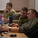 Three military personnel in camouflage and flight suits are working at a desk with laptops and computers in what appears to be a briefing room or operations center. Two individuals in the foreground are collaborating closely, looking at a shared screen, while a third service member works independently in the background. The setting suggests a training exercise, mission planning session, or operational briefing environment.