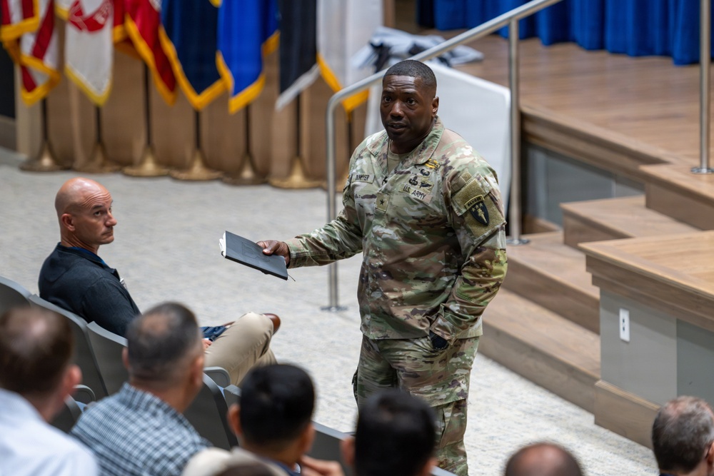A U.S. Army soldier in OCP (Operational Camouflage Pattern) uniform is standing in what appears to be an auditorium or briefing hall, holding a small notebook or tablet. He appears to be addressing or interacting with an audience seated in rows. The setting suggests a formal military briefing, ceremony, or training event.