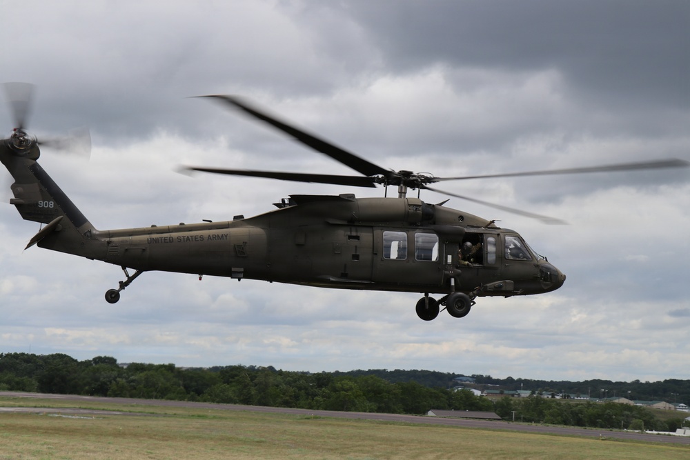 A United States Army UH-60 Black Hawk helicopter, tail number 908, is captured in low-altitude flight over what appears to be an airfield or military installation. The helicopter is painted in olive drab/army green and bears clear 'UNITED STATES ARMY' markings on its fuselage. At least one crew member is visible through the cabin windows, and the main rotor blades show motion blur indicating active flight.